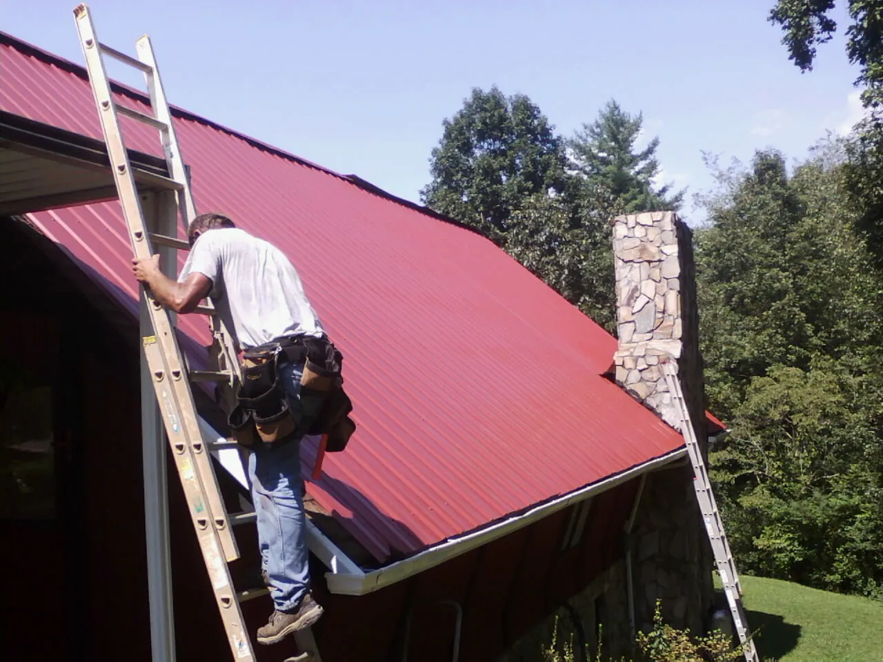 Roofer on a ladder beside a red metal roof and stone chimney — Soffit & Fascia Repair by Roofing Brothers in Chino Hills
